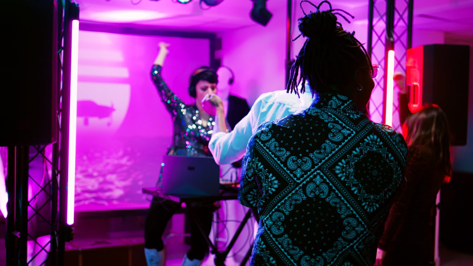 Young adult putting music at party, working as DJ at discotheque with friends. Woman using audio mixing station on stage with discolights, having fun with group of people. Handheld shot.